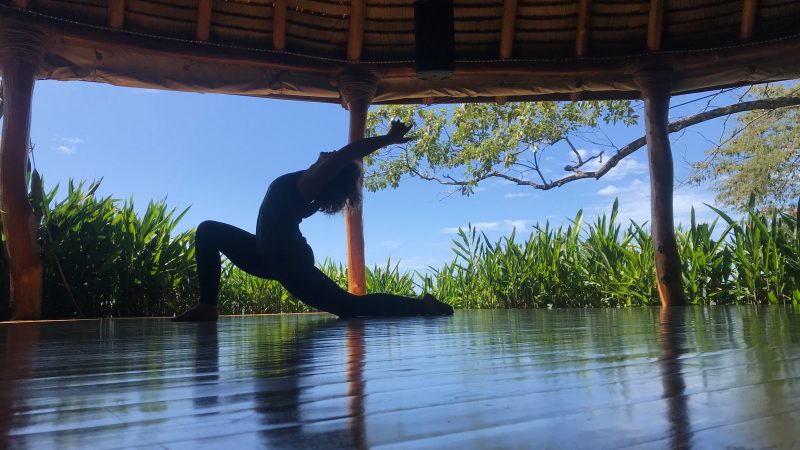 The silhouette of a women practicing yoga in a large pavilion. Trees and blue sky in the background.