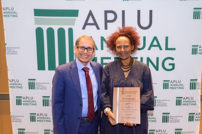 A man and woman pose for a photo in front of an APLU banner.