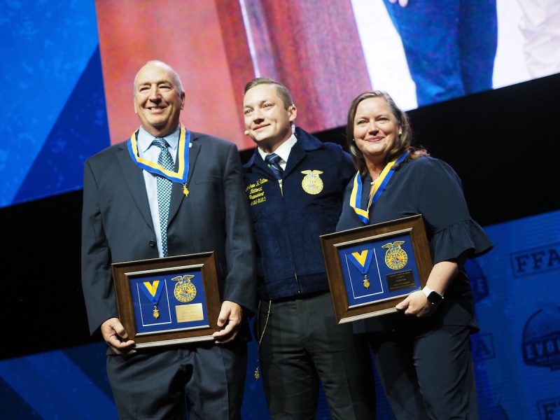 Two men and a woman stand on a stage, brandishing FFA awards and smiling.