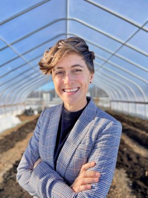 white woman with short hair in blue suite smiling, arms crossed in front of her chest, standing in a greenhouse.