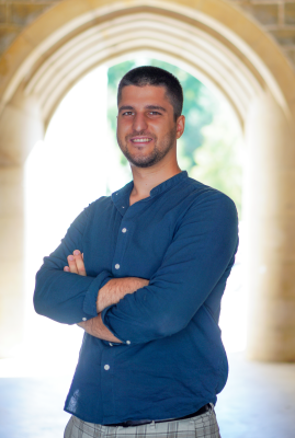 Man with light skin and brown hair is wearing a blue button-up shirt standing under an archway. His arms are crossed in front of his chest and he is smiling. 