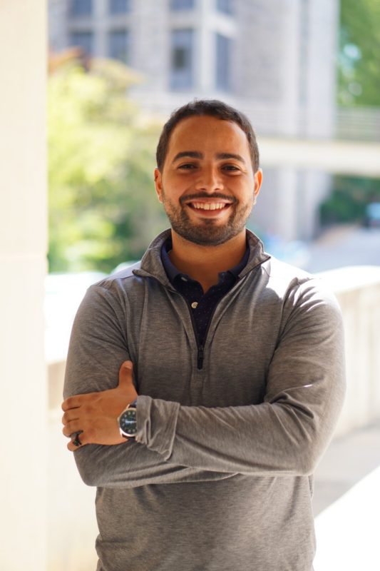 Man with tan skin standing outside with blurred background, crossing his arms in front of his chest; smiling