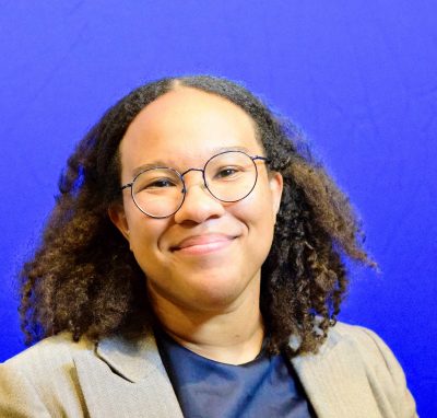 Woman with tan shin and medium length curly hair wearing a blue shirt and tan blazer; smiling and wearing glasses