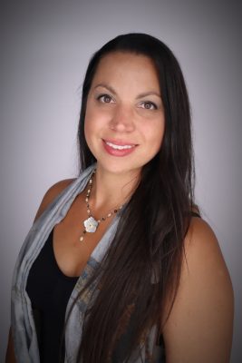 Woman with tan shin and long black hair smiling in front of a grey background. 