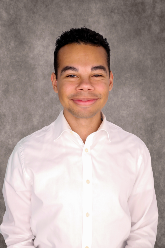 Man with tan skin and short black curly hair wearing a light button up shirt. 