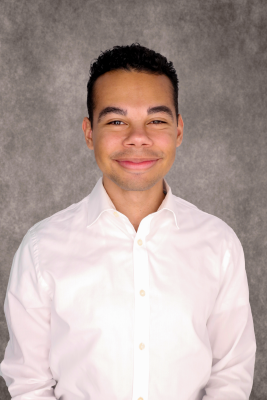 Man with tan skin and short black curly hair wearing a light button up shirt. 
