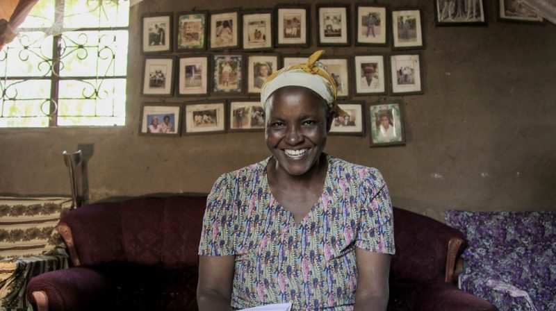 A smiling woman sits on a couch in front of a wall filled with photographs 