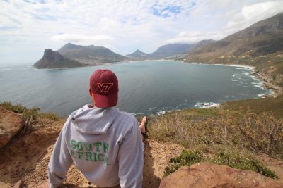 A student wearing a Virginia Tech hat looks off at the lake and mountains in the distance.