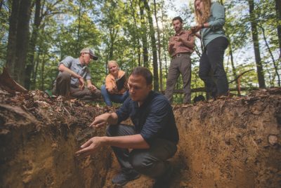 A person kneels in a hole in the ground to take a sample of soil, as three other people look on.